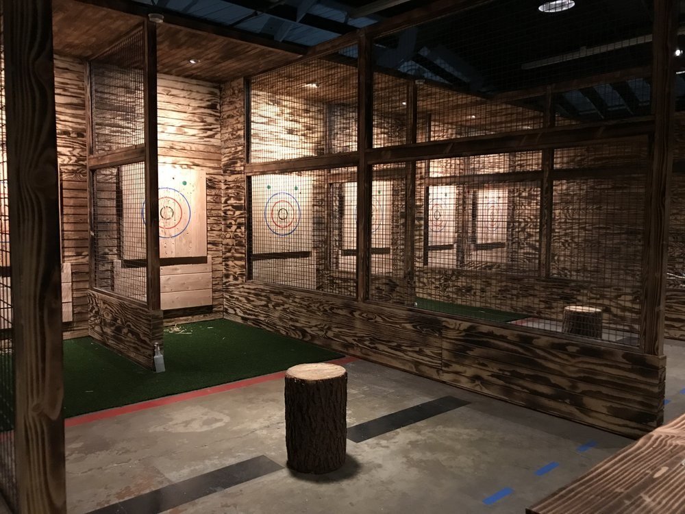 The throwing floor at AXT in Costa Mesa: caged lane bays with burnt wood walls, a painted target at the end of each lane, green turf under the targets and a stump at the throwing line. Source: a guest's photo on the venue's public Yelp listing.